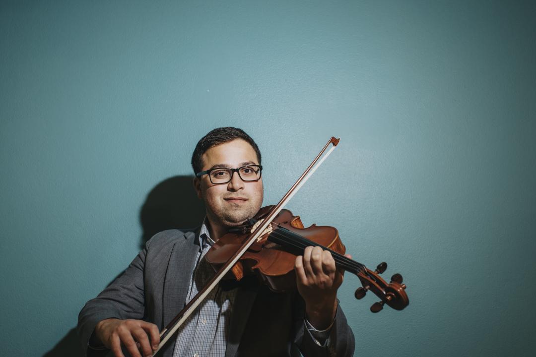 Seen from the lower torso up, and standing against a blue background facing the camera, Jon Torre plays his violin.
