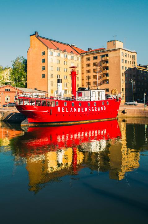 Viewed from the katajanokka shore, the bright red lighthouse ship Relandersgrund floats in front of an apartment block. The sky is blue and Relandersgrund reflects clearly in the water.  