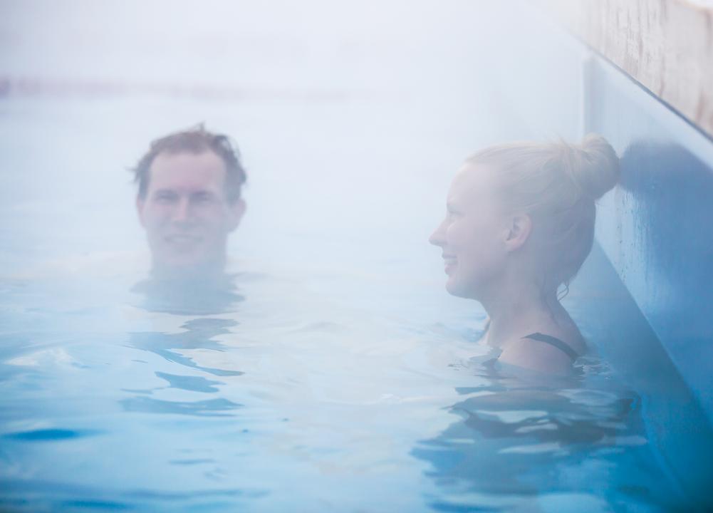 Showing mainly the water, a man facing the camera and a woman to the right resting against the wall smile, as they show their heads above the water at a misty Allas Sea Pool
