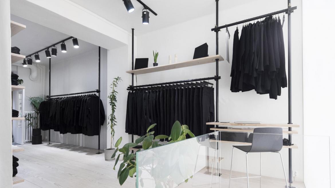 In a brilliant white store interior, a series of three black clothes on racks against the far wall are filled with various black clothing.