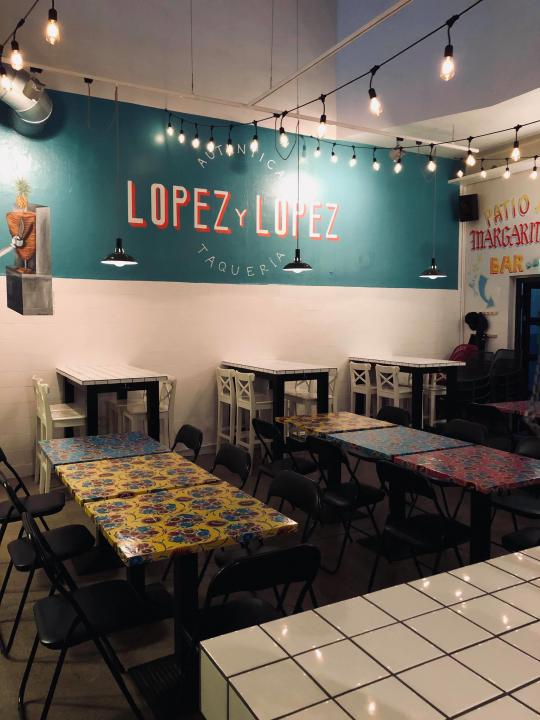 Inside a Lopez y Lopez Mexican restaurant, a white, square tiled service counter can just be seen on the bottom right of the photo and a large blue, banner with the restaurant logo sits on the opposite wall. Between these and beneath rows of rope lights, are three, four-seater tables and six, two-seater tables.