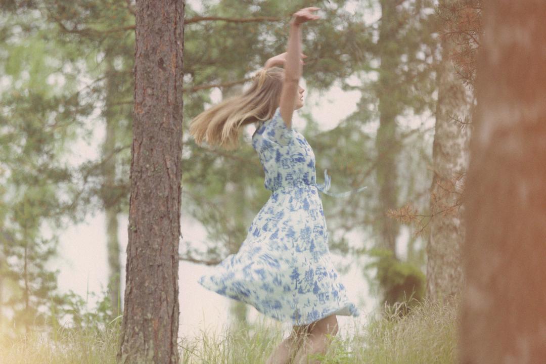 A woman in a summer dress is spinning around in the woods with her hands above her head.