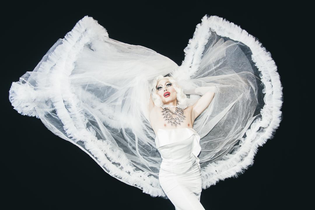 Betty Fvck strikes a pose, hands above their head,in white gown against black background.