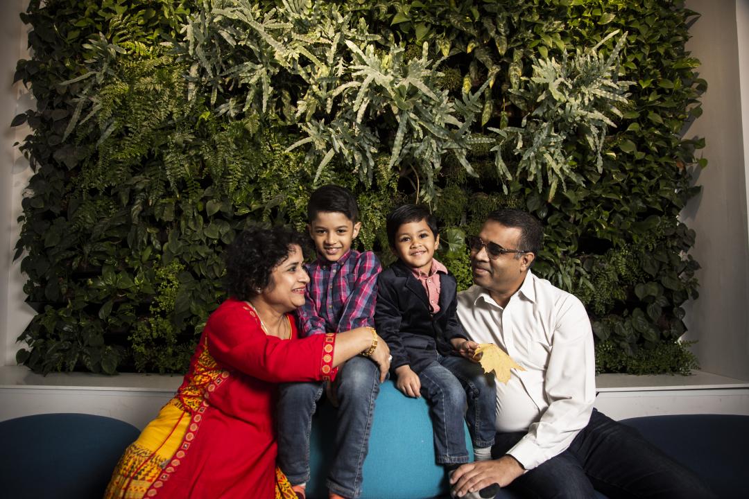 Father and mother sit with their two children between them, looking into each other's eyes, kids smiling at the camera. The wall behind them is filled with live, green plants.