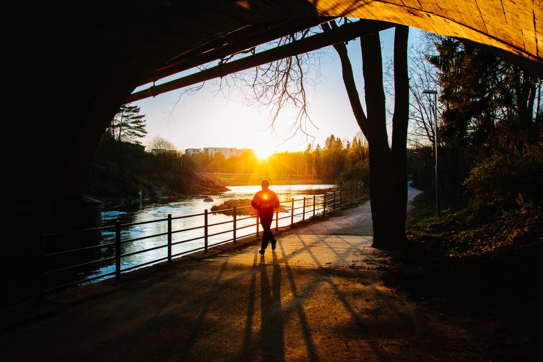 A person takes an evening run by Vanhakaupunginlahti bay. They're passing underneath a stone bridge with the river and, a sun that has almost set to their back, casting long shadows towards the camera.