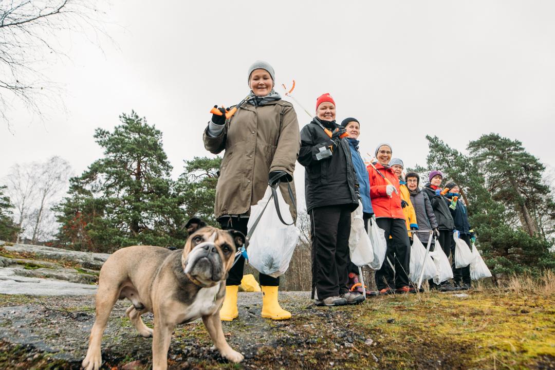 7 people who have volunteered to clean the sea shores, stand in a line leading away from the camera to the right on a rocky slope, a few trees in the background. They all carry white bin bags, smiling at the camera, while a bulldog stands in front of the line, looking towards the camera.