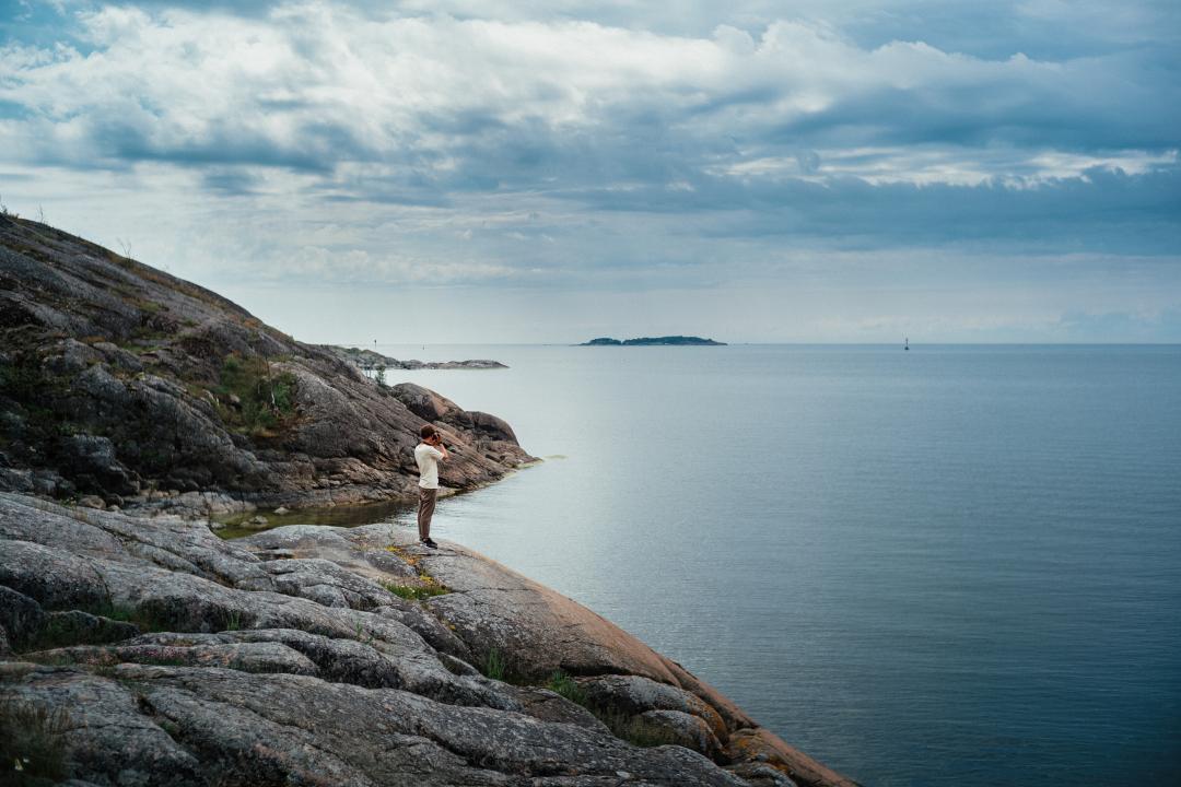 In the distance, a man stands by the sea to take a photo in the Helsinki archipelago, the open sea to his right, the rocky shore rising gradually to his left. Stormy looking clouds are building in the sky.