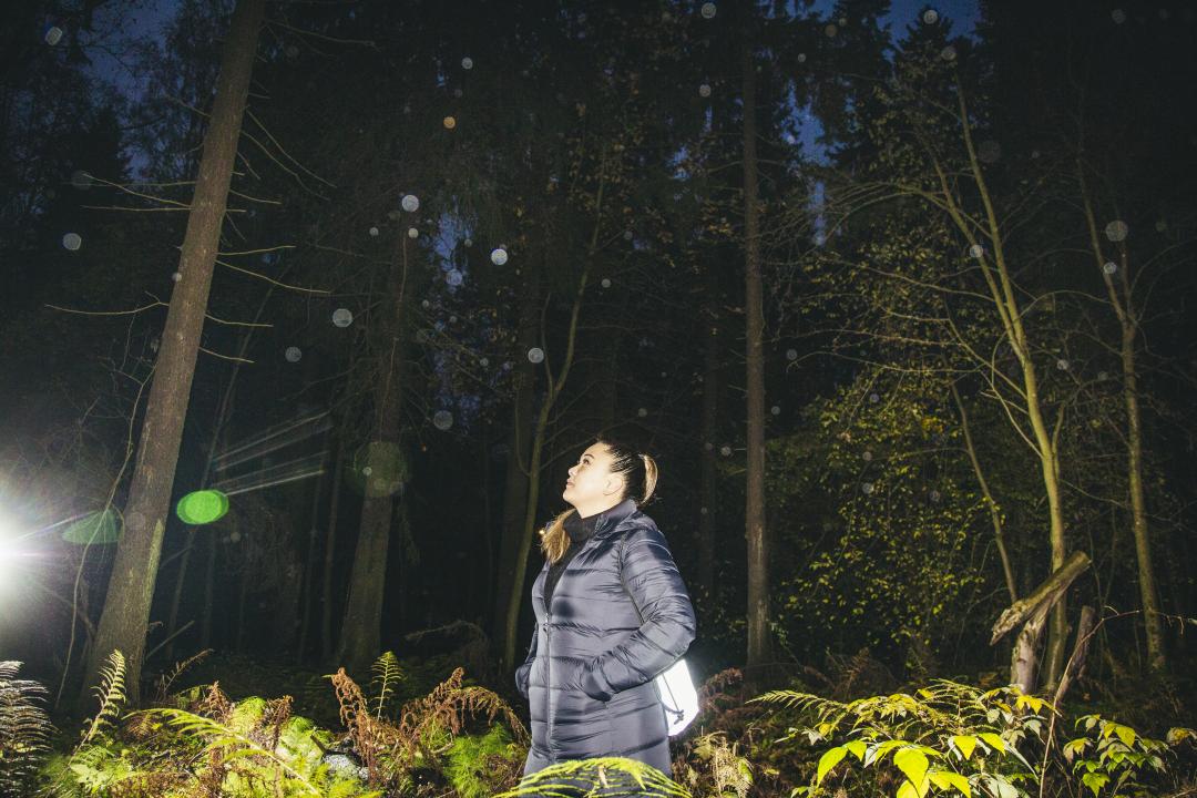 Adele Asuncion standing in the woods in the dark, looking up into the tree tops to the left.