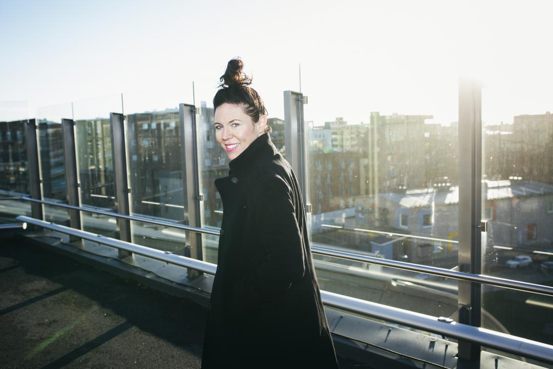Mary Morgan walking away from the camera on Helsinki rooftop, turns and smiles, the Helsinki cityscape behind her.