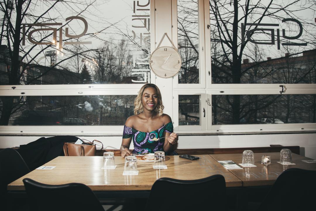 Mariam Shodeinde sits alone at a long table in a restaurant, facing the camera and smiling, large windows showing an elevated view above the city behind her.