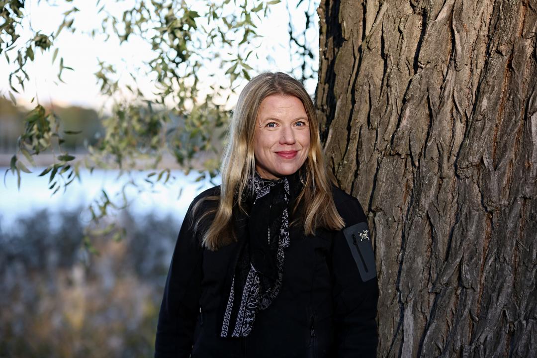 Portrait of Salla Tuomivaara, looking at the camera smiling, she is standing next to a large tree .