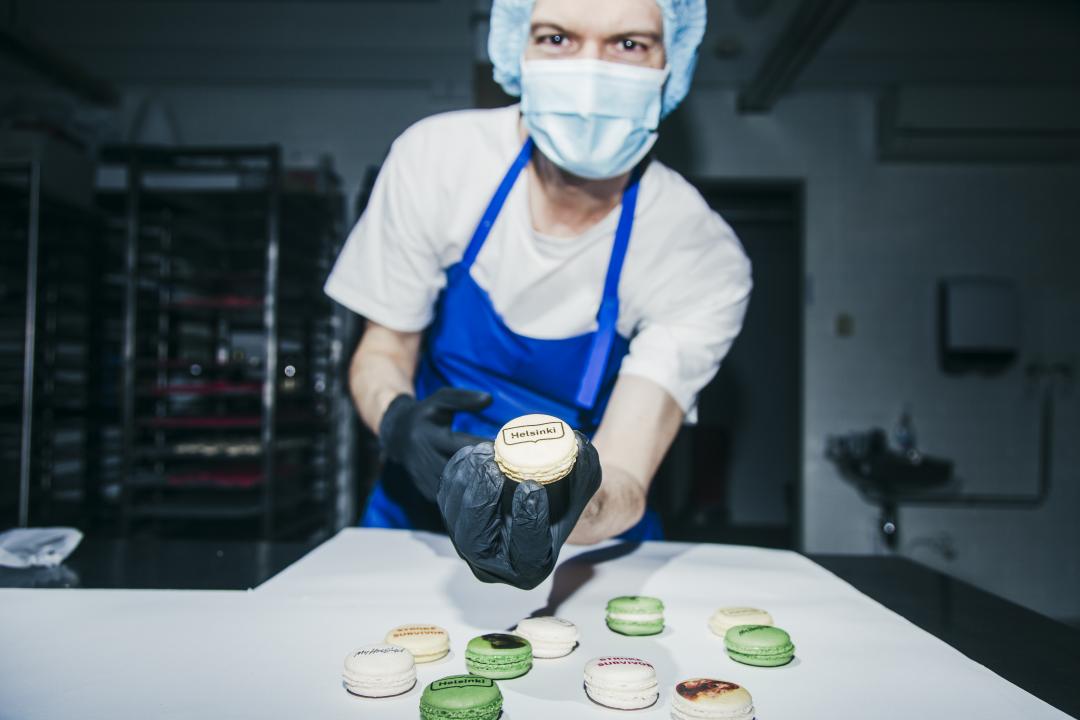 Olivier Béaslas dressed in a blue apron, hair net and wearing a face mask is standing over a work surface in a professional kitchen showing a macaroon he's made to the camera. More macaroons arranged on the work surface.