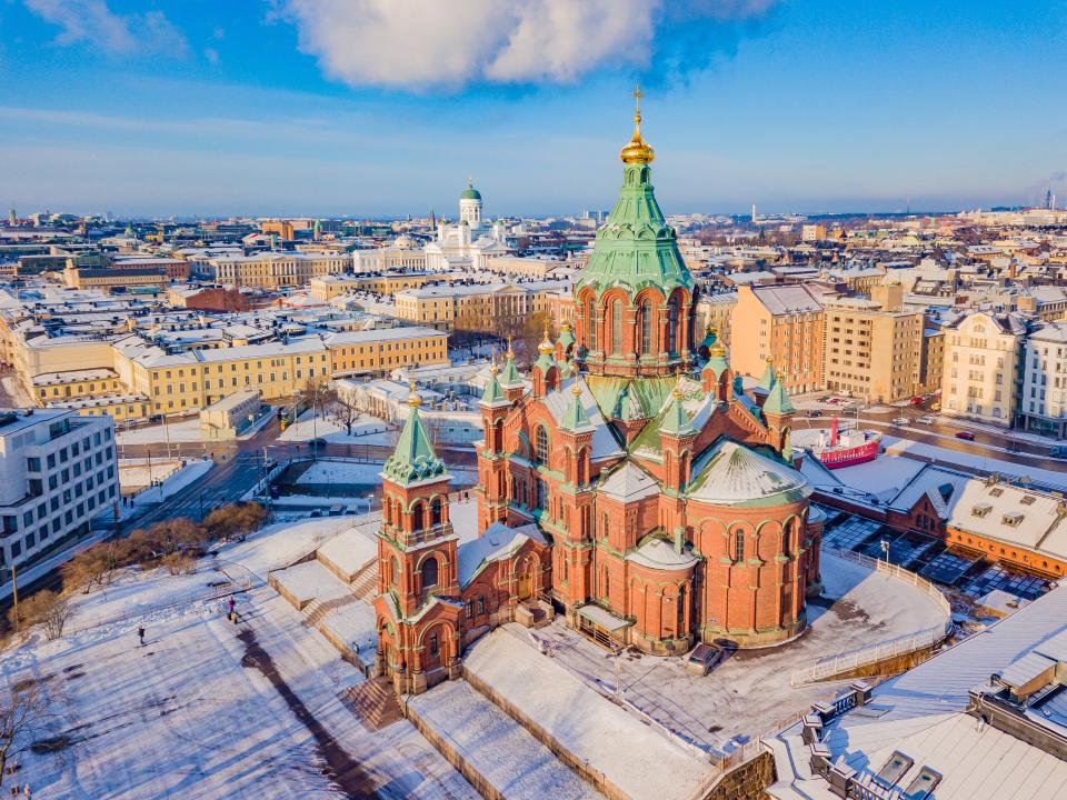 An aerial view of Uspenski Cathedral in winter, the Helsinki cityscape stretching into into the horizon, Helsinki Cathedral standing out from amongst the skyline in the distance. Everything has been lightly covered by snow.