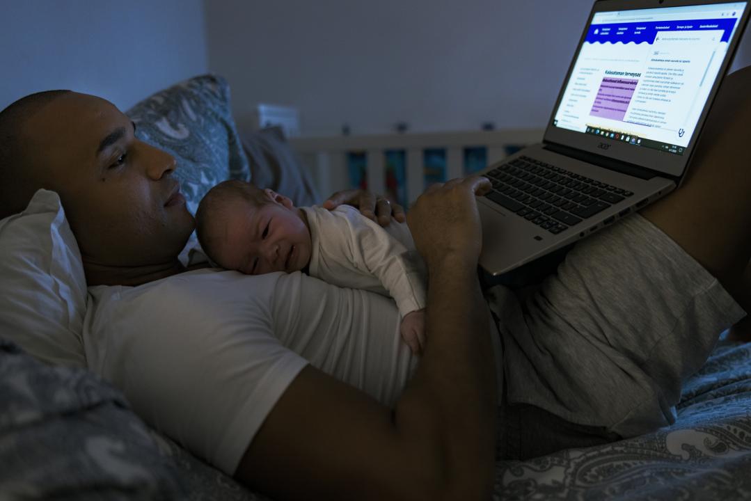 Lying down together, a father and baby chill at home. Baby lies on the father's chest while the father balances an open laptop on his lap. 