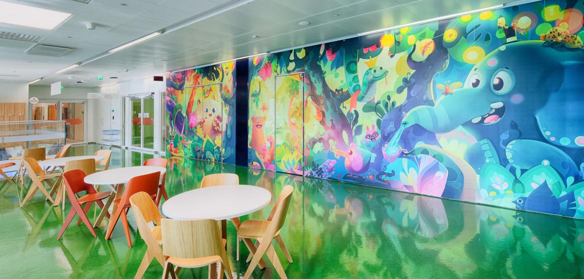 In a long room with a green floor and holding 3 circular tables, each with 4 chairs, a huge and colourful mural of animals in the jungle covers the far wall.