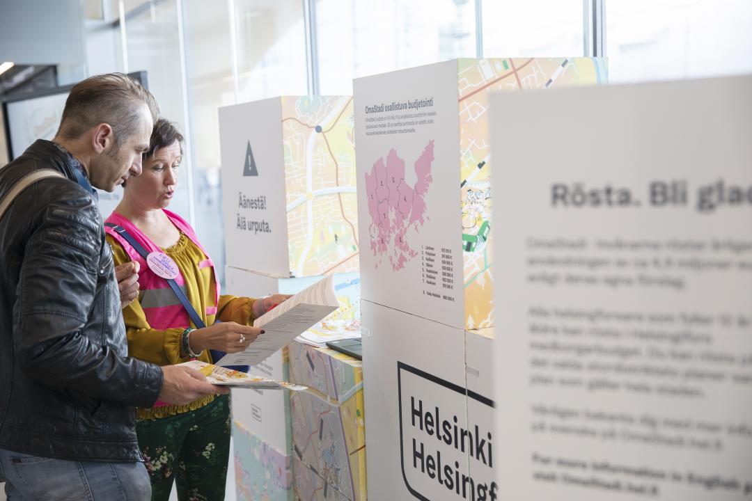 Two people at an Omastadi workshop look at information leaflets and discuss in front of a Helsinki City information stand.