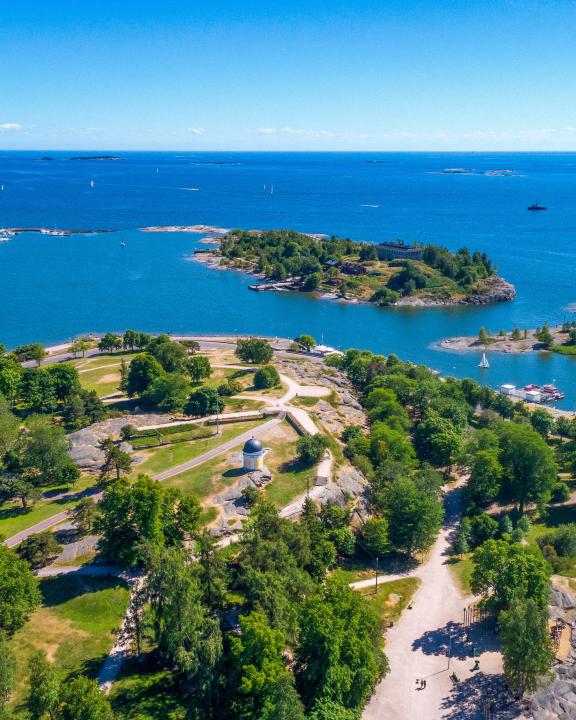 Aerial of Kaivopuisto park full of green trees surrounding the observatory at its peak, overlooking a magnificent view of a bright blue sea stretching into the horizon on a sunny day.