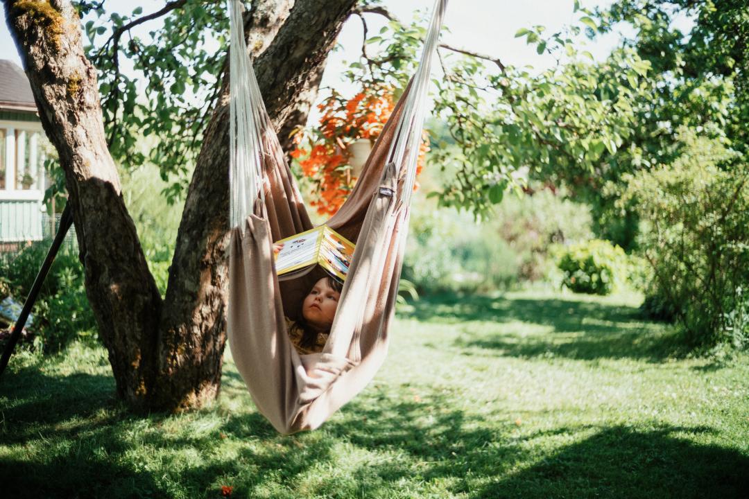 A kid is relaxing in a pink hammock, hanging from a tall v-shaped tree located in a green and grassy allotment garden