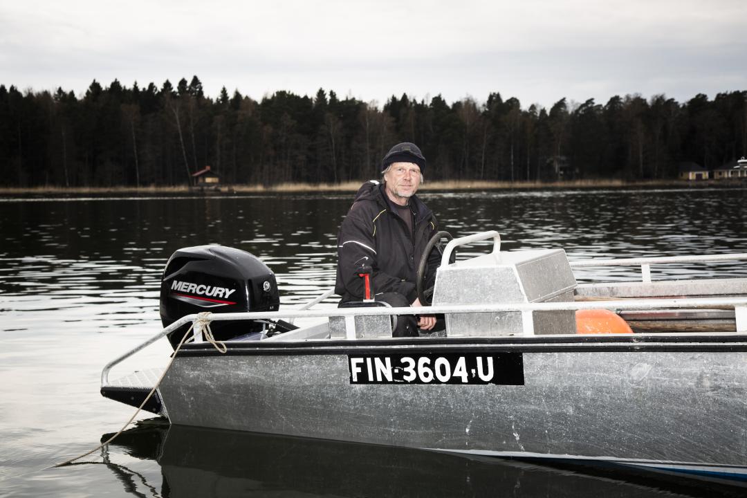 Kai Ilves sitting in a small motorboat on calm water, where the dark outline of the forest can be seen running along the shore in the distance. 