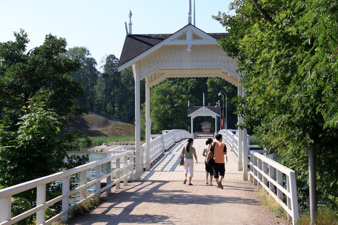 Three people walk under the white, wooden archway across an empty Seurasaari bridge, trees in full bloom at the entrance to the bridge and on Seurasaari beyond.