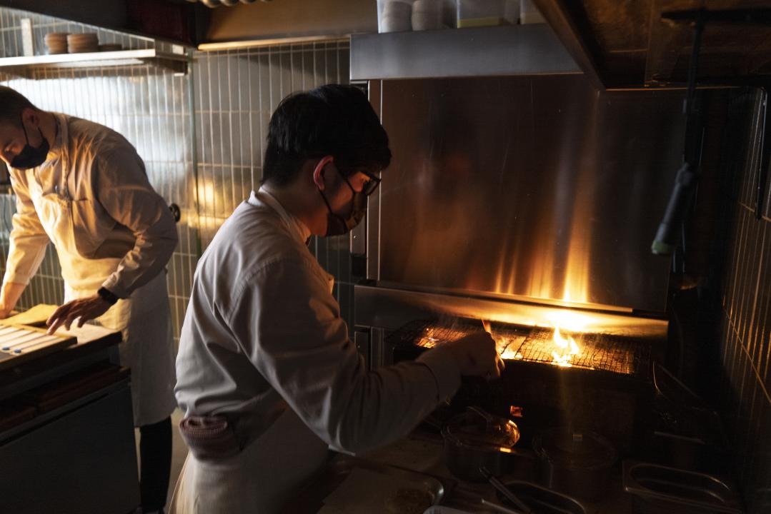 Looking over a lit stove, a chef prepares food over an open fire at Restaurant Grön, while a colleague is busy in the background checking something to the left. 