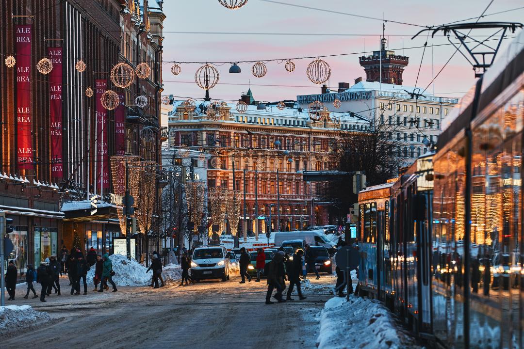 A wintry looking scene on Mannerheimintie, with Christmas lights suspended above the street and a pink and blue sky in the background. A tram is waiting for people to cross the street ahead, with Stockmann department store standing on the left  hand side.