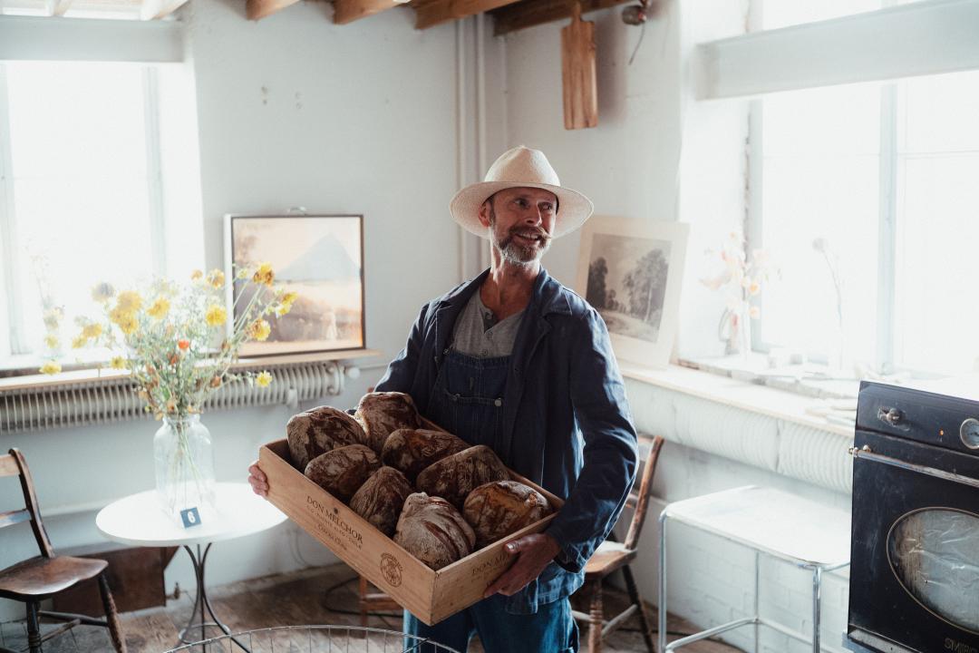 Male baker inside Bakery Ambience at the Venetsia Building, smiling whilst holding a large box of crusty loaves