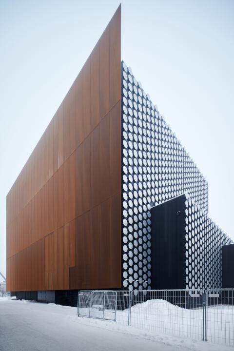 External view of the tall Dance House Helsinki building, as seen from the corner. The left side of the building is covered in rusted panels whereas the right hand side is covered with small silver discs.