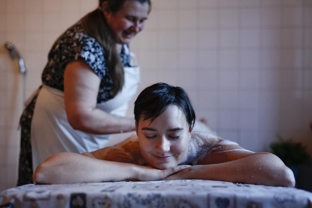 Girl lying down on a table, facing the camera with their arms folded under their chin, whilst a lady washes them at Kotiharjun sauna.
