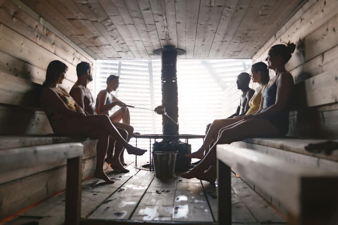 Bunch of people sitting in wooden sauna, silhouettes visible and bright light shining through from the background window