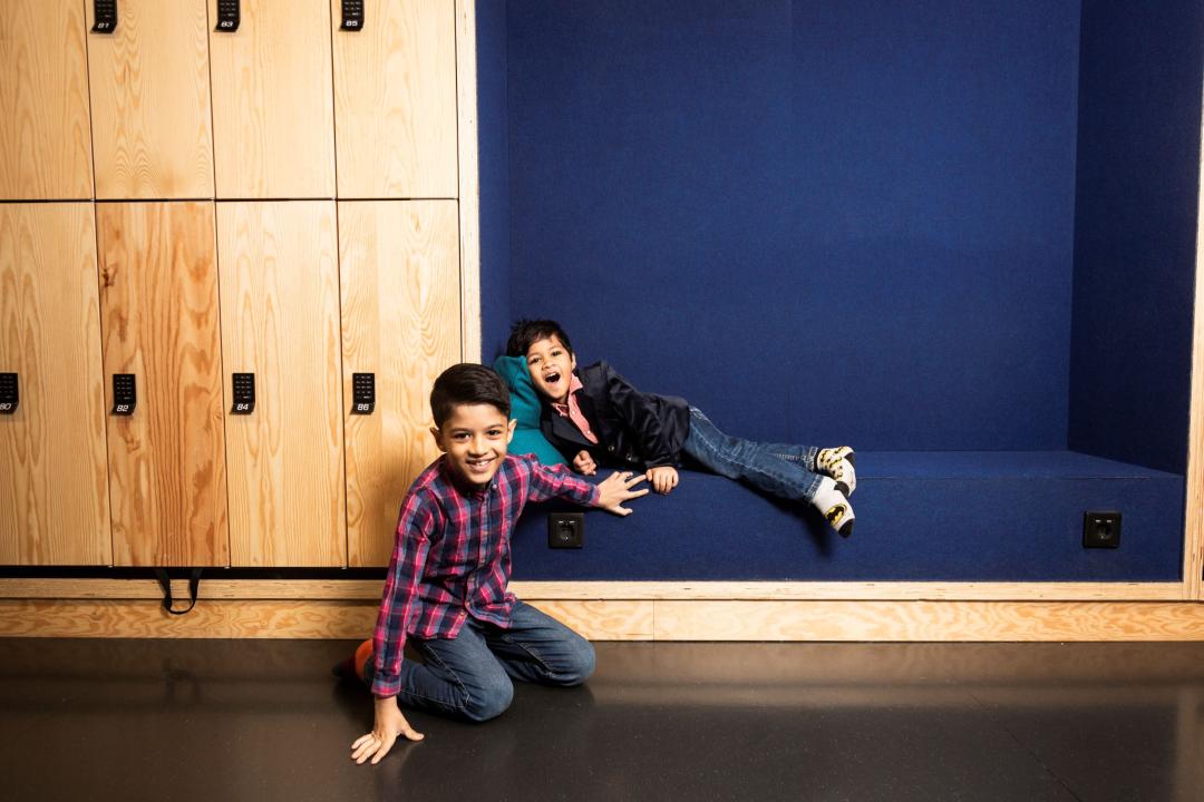 The two boys, Gaurang and Shivam, pose for a photo against wooden clad lockers and on a blue bench. One kneeling on the floor, the other lying on the bench, both are smiling.