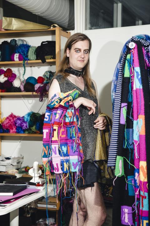Juha Vehmaanperä standing and holding a colourful fabric, more colourful fabrics on the right side, balls of yarn on shelf on the left side