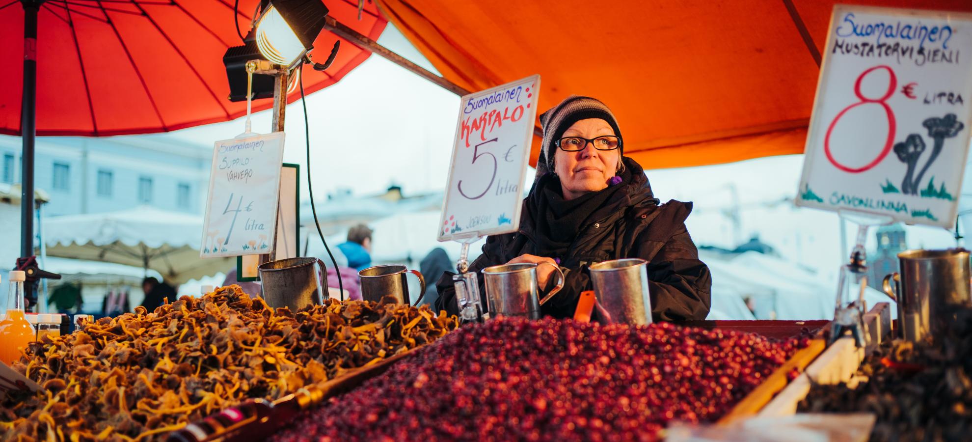 Underneath an orange market stall canopy, a woman dressed in a black coat, hat and looking to the right of the photo stands behind her sales counter, which is raised at a slight angle, and is packed full of cranberries and Chanterelle mushrooms. 