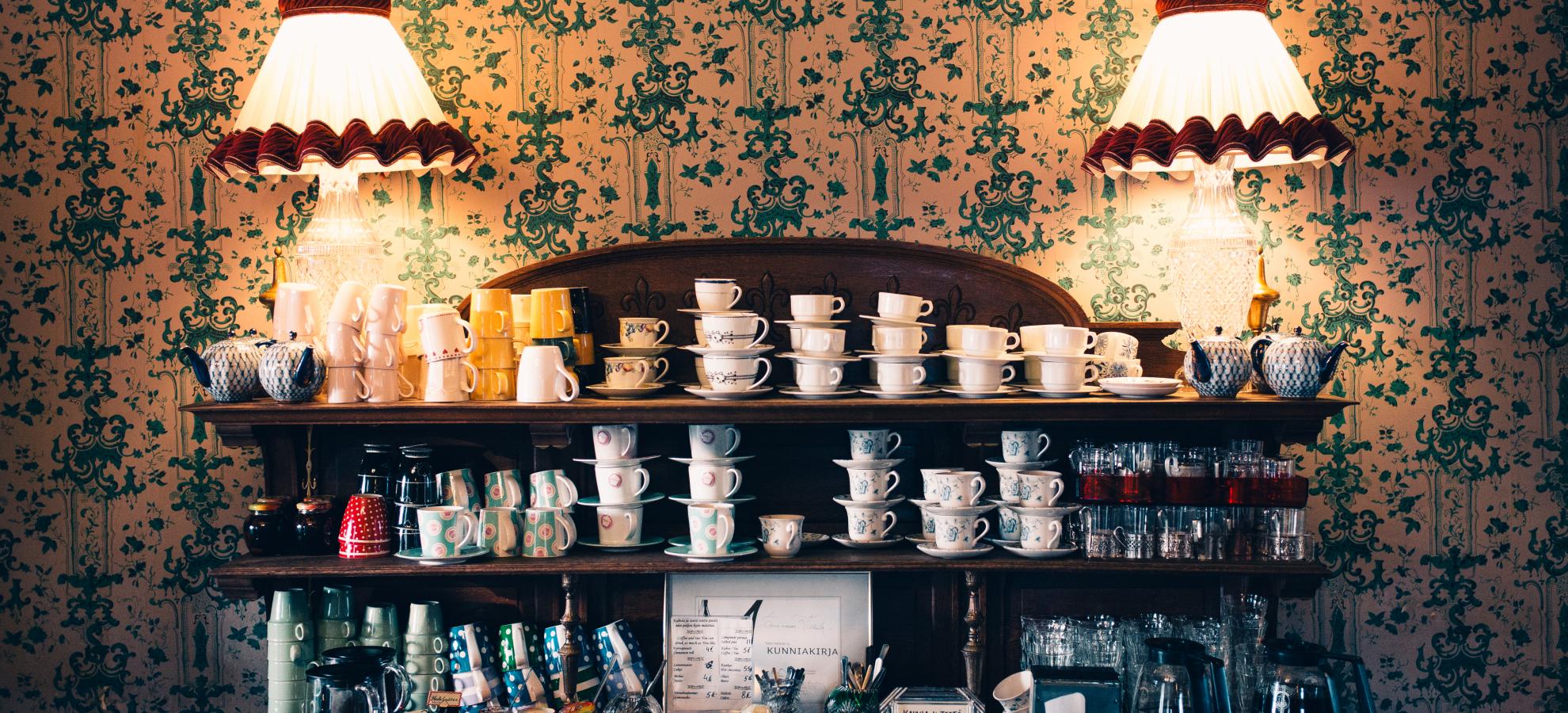 In front of a heavily patterned, cream and green traditional wallpaper, stands two lamps either side of a mahogany cabinet, its shelves full of teacups, mugs and glasses. 
