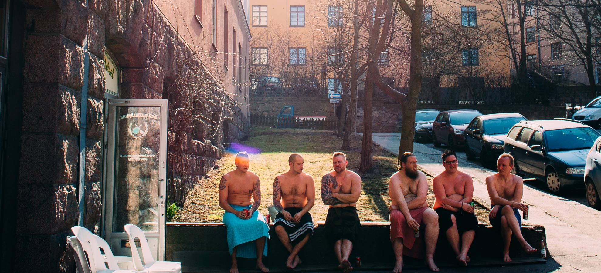 Six men in towels sit on the wall cooling off outside of Kotiharju sauna on a chilly, sunny day.