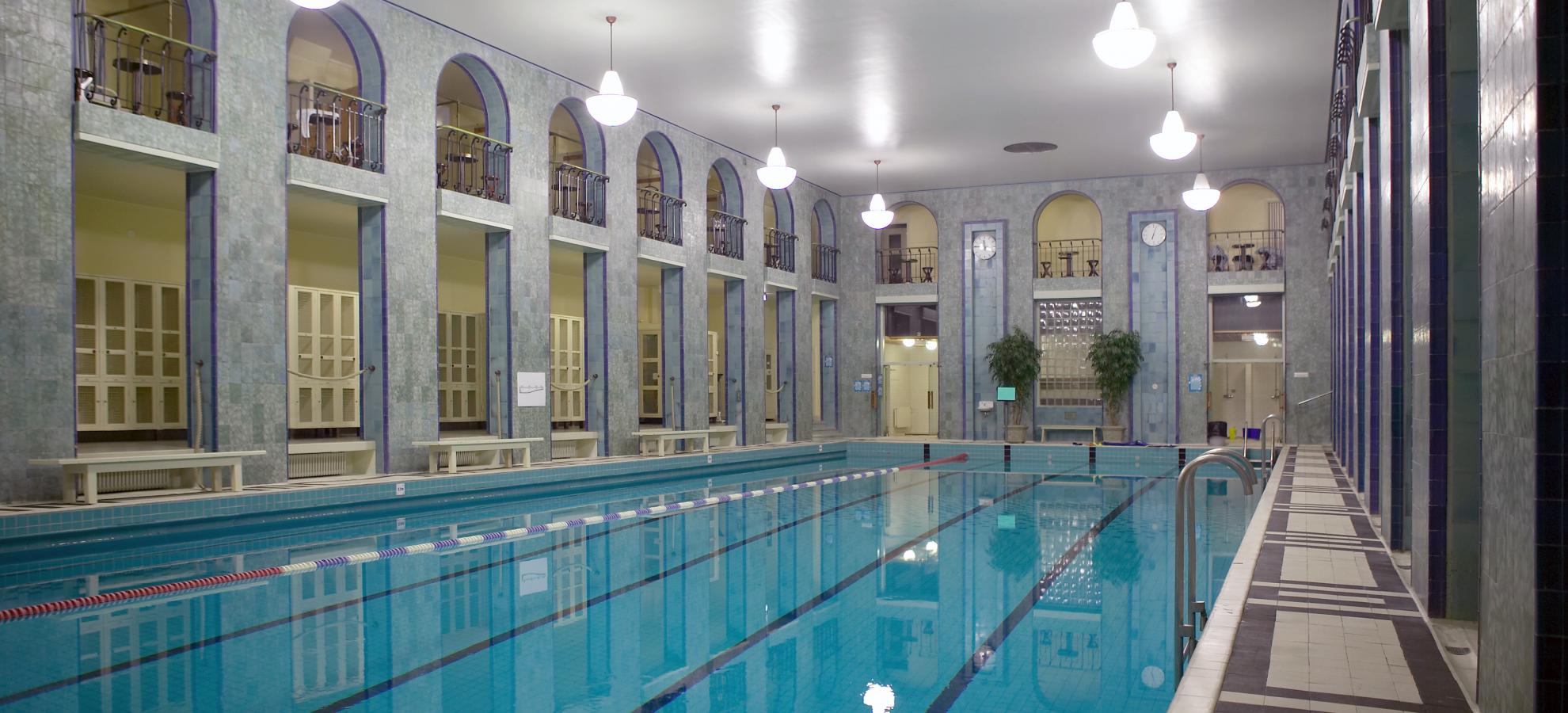Inside Yrjönkatu's swimming hall, the long pool stretches many metres to the back of a grand hall surrounded by multiple archways, with two rows of chandelier-like lights hanging above the pool.