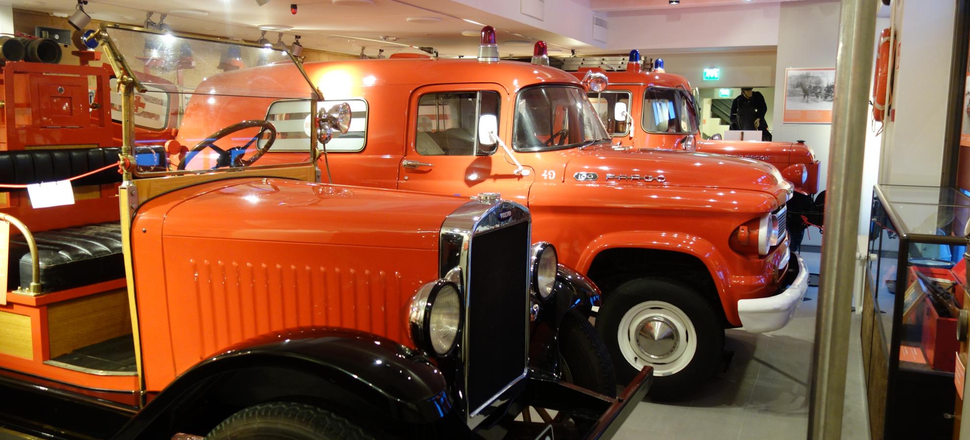 Inside one of the large exhibition spaces of Helsinki City Rescue Department's Fire Museum, a row of three old, orange service cars and vans from different ages stand in a row leading away from the camera, almost filling the photo.