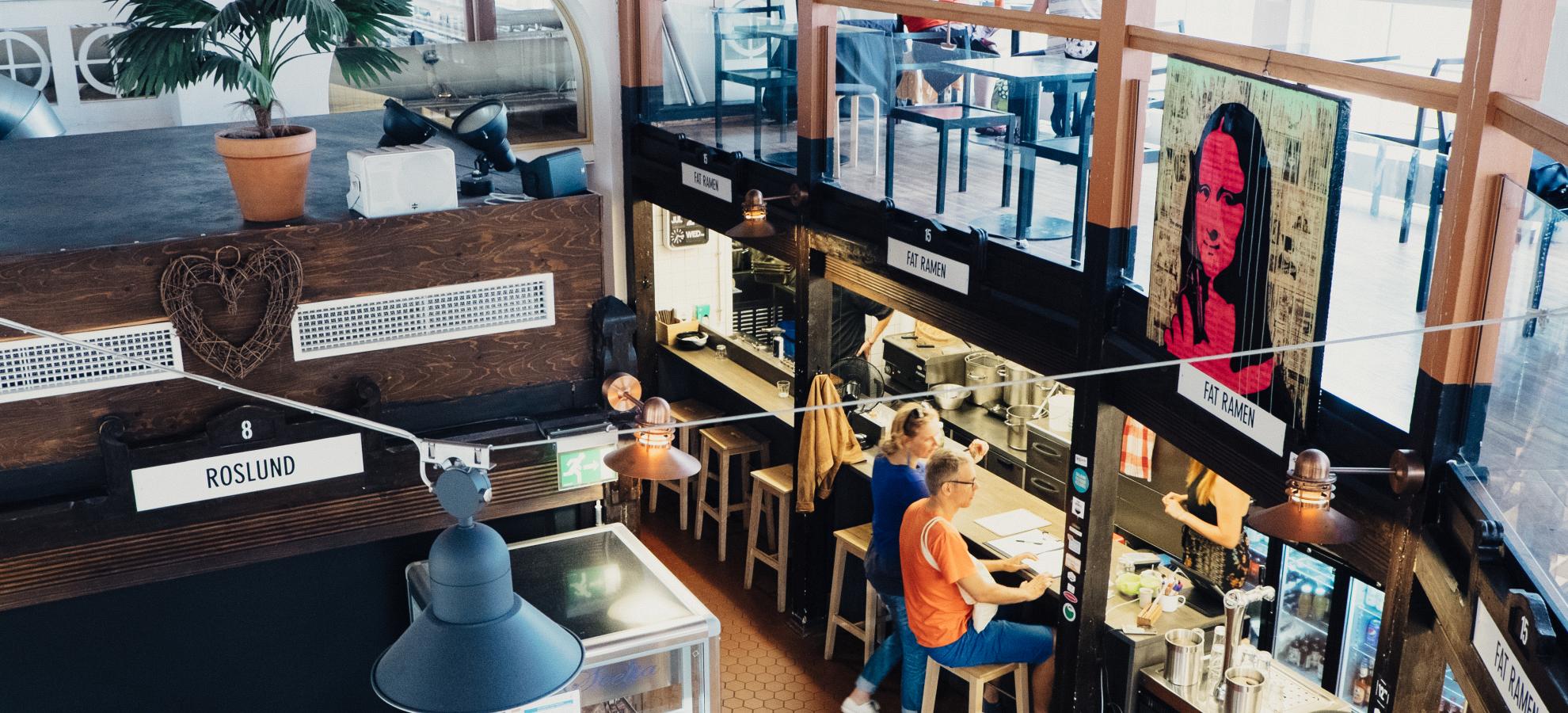 A view from above inside Hietalahti's Market Hall. Below customers sit on high stools, eating at the bar of a Ramen restaurant, whilst other customers sit in an open seating area directly above the restaurant itself. 
