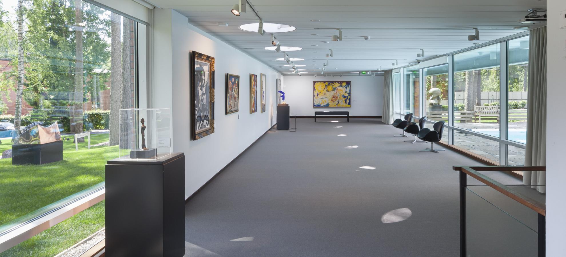 A long exhibition space inside Didrichsen Art Museum, large windows flank either side of an almost empty room, the left and far walls holding a handful of larger paintings together. 