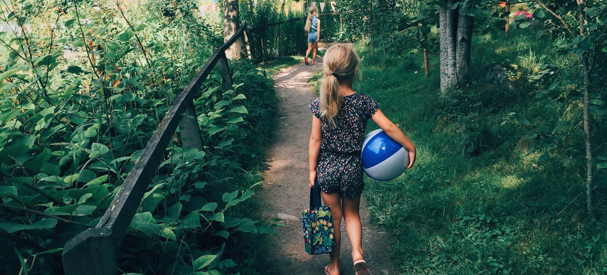 Facing away from the camera, a little girl carrying a beach ball under one arm and a bag in her opposite hand, walks down a trail that is surrounded by grass, bushes and trees.
