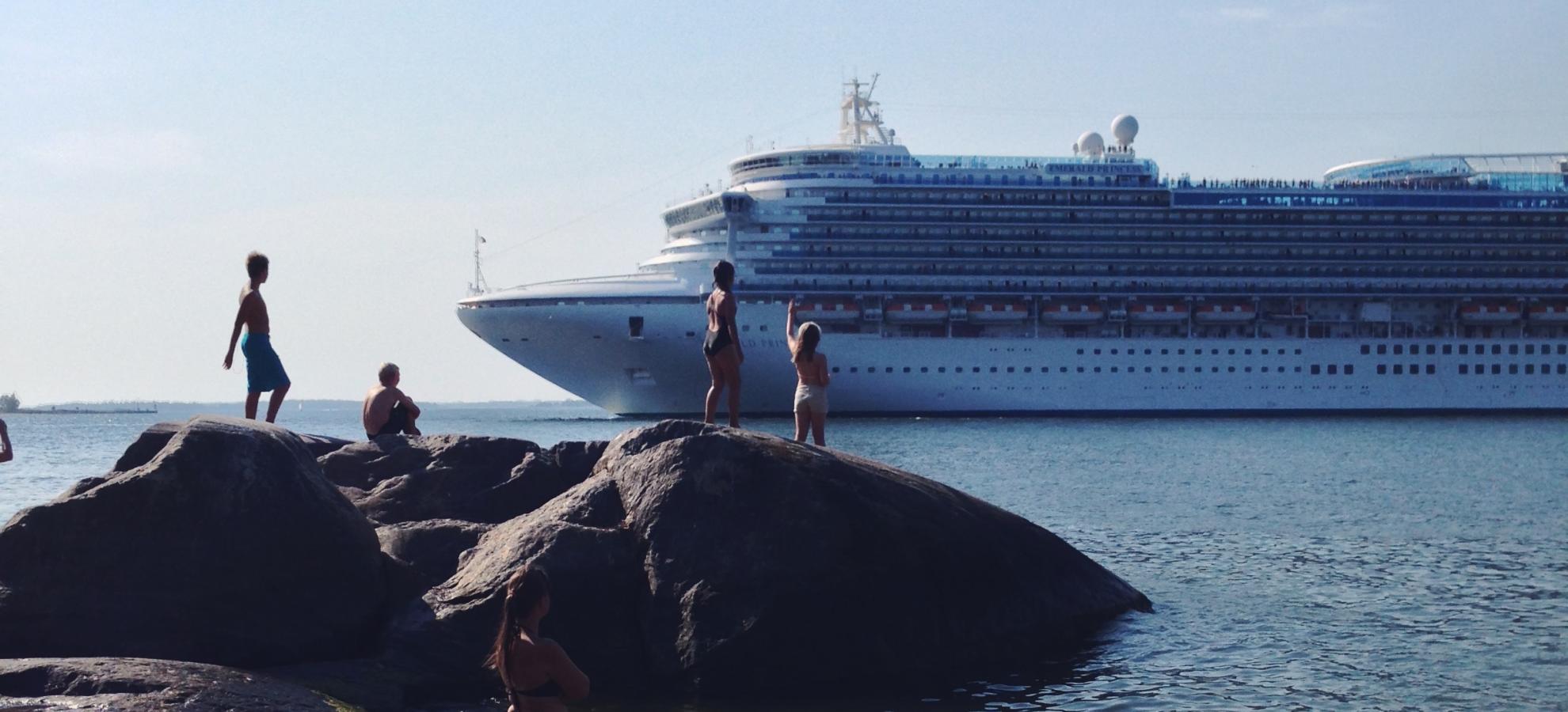 As a cruise ship passes by in the distance, a small group of sunbathers and swimmers stand in the water and on nearby rocks, watching and waving at the ship.