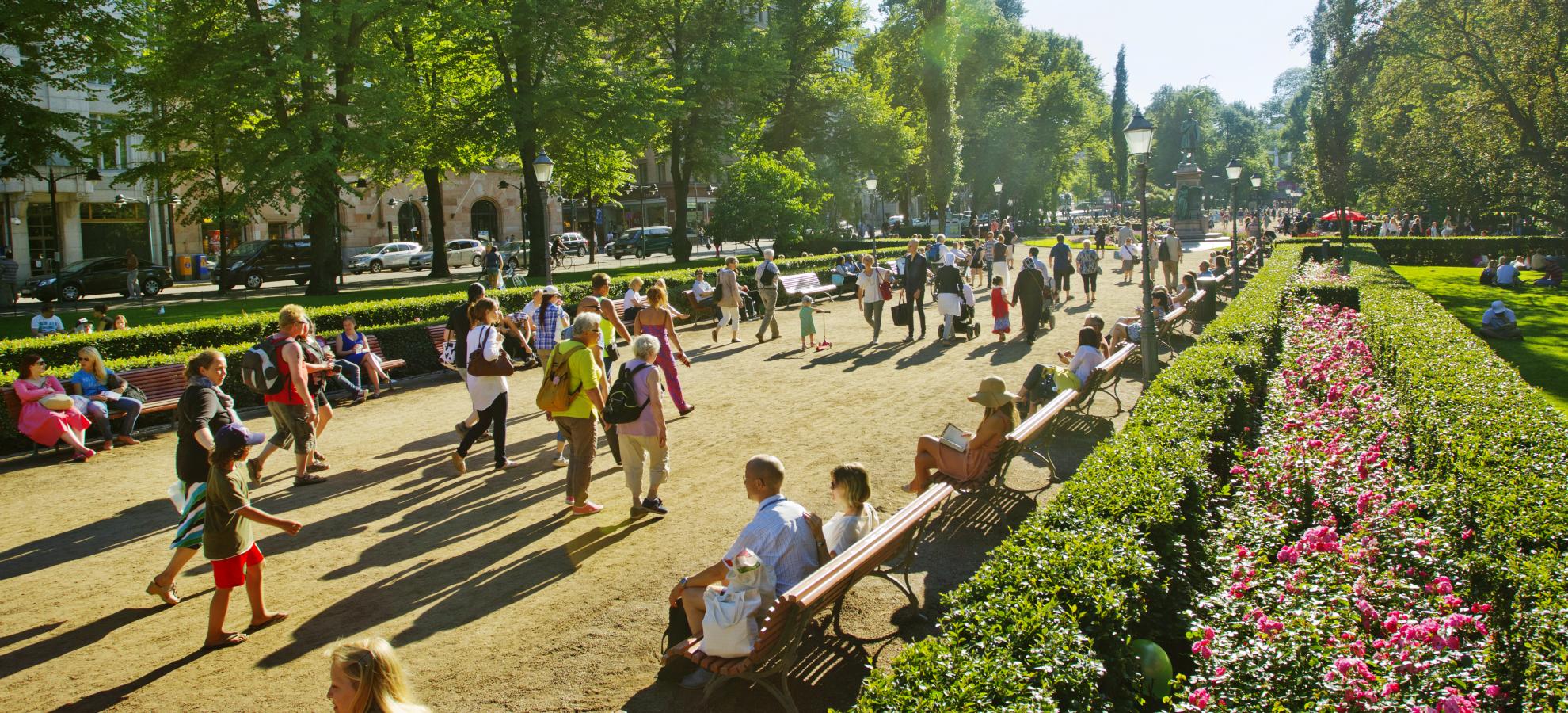 A view looking down the length of part of Esplanadi park in summertime. Surrounded by neat bushes, flower beds and trees, crowds of people walk up and down the main walkway.  