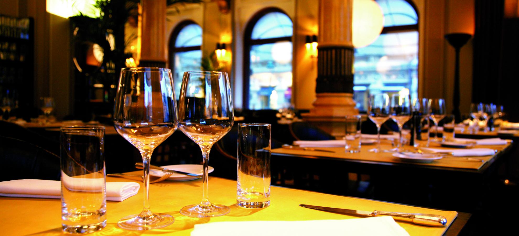 Inside Brasserie Kämp, dozens of tables have been set with fine glassware, tall windows on the far side of the room with columns standing in front of them. The lighting is gentle and looks golden.