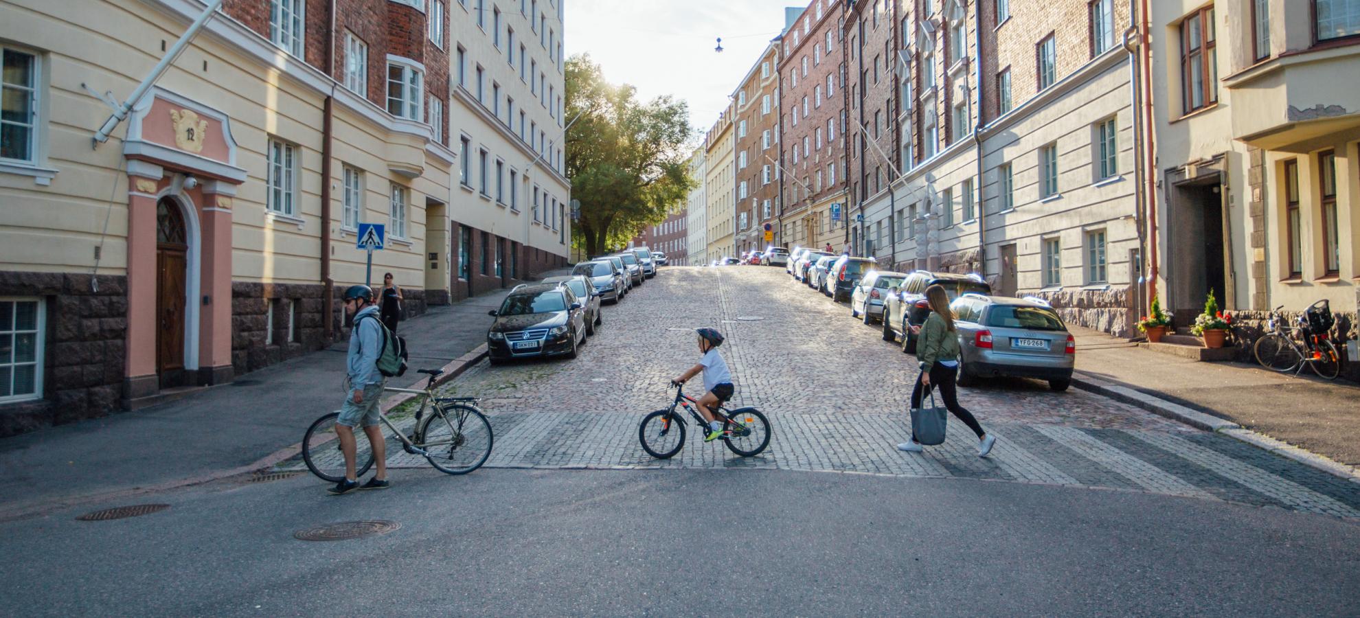 Cyclists and a pedestrian cross a street crossing in Töölö, with a variety of traditional apartment buildings going up the hill either side.