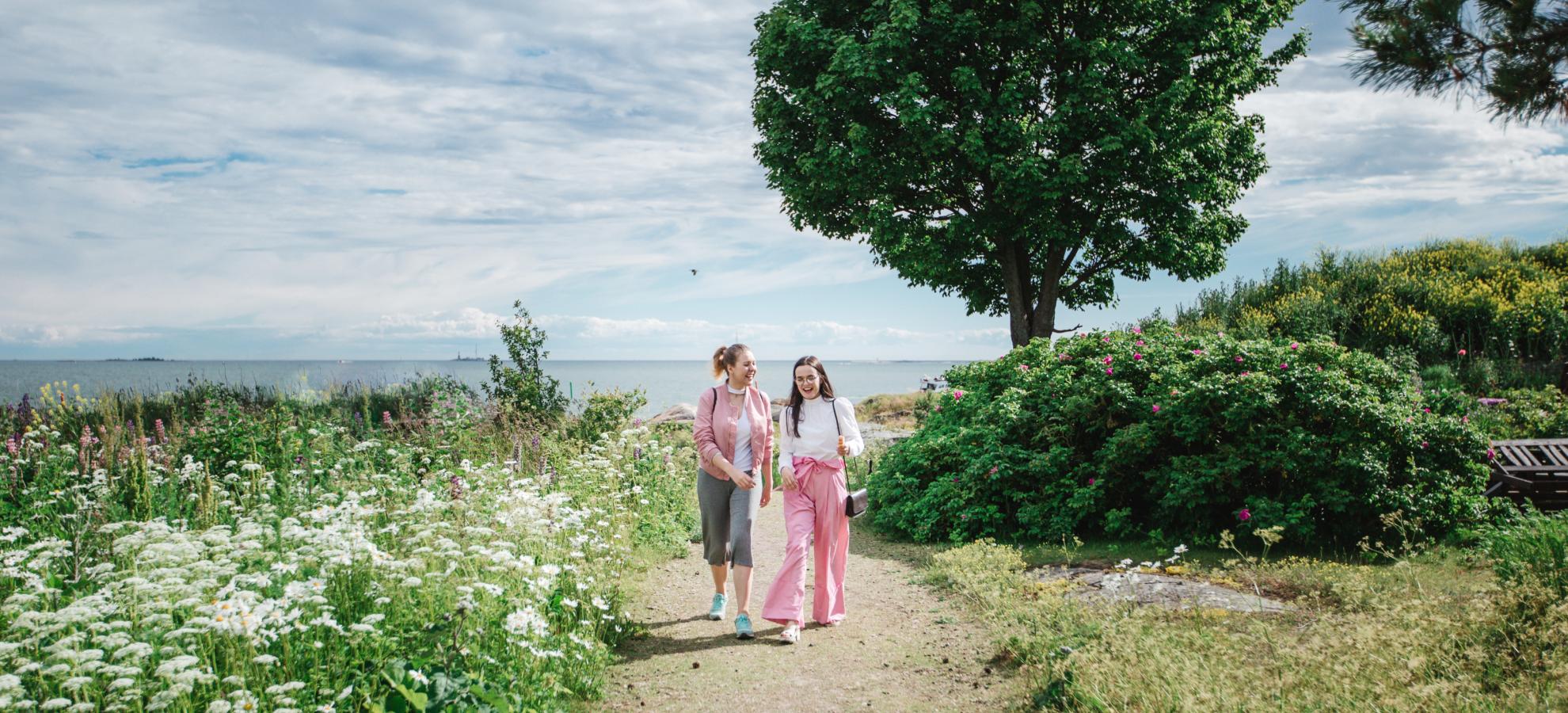 Walking towards the camera, two girls stroll along a path on Suomenlinna during a sunny day. Either side of the path are wildflowers, bushes and a tree, with the sea just seen on the horizon.