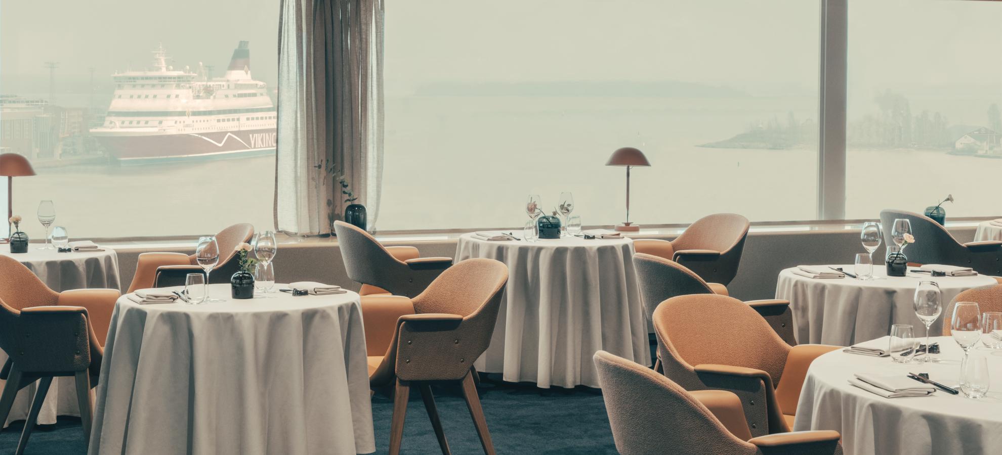 A view from inside Ravintola Palace overlooking a very misty Helsinki harbour, a cruise ship docked to the left. 5 tables can be seen in the photo, neatly set with folded, white table cloths with 70's style designer chairs.