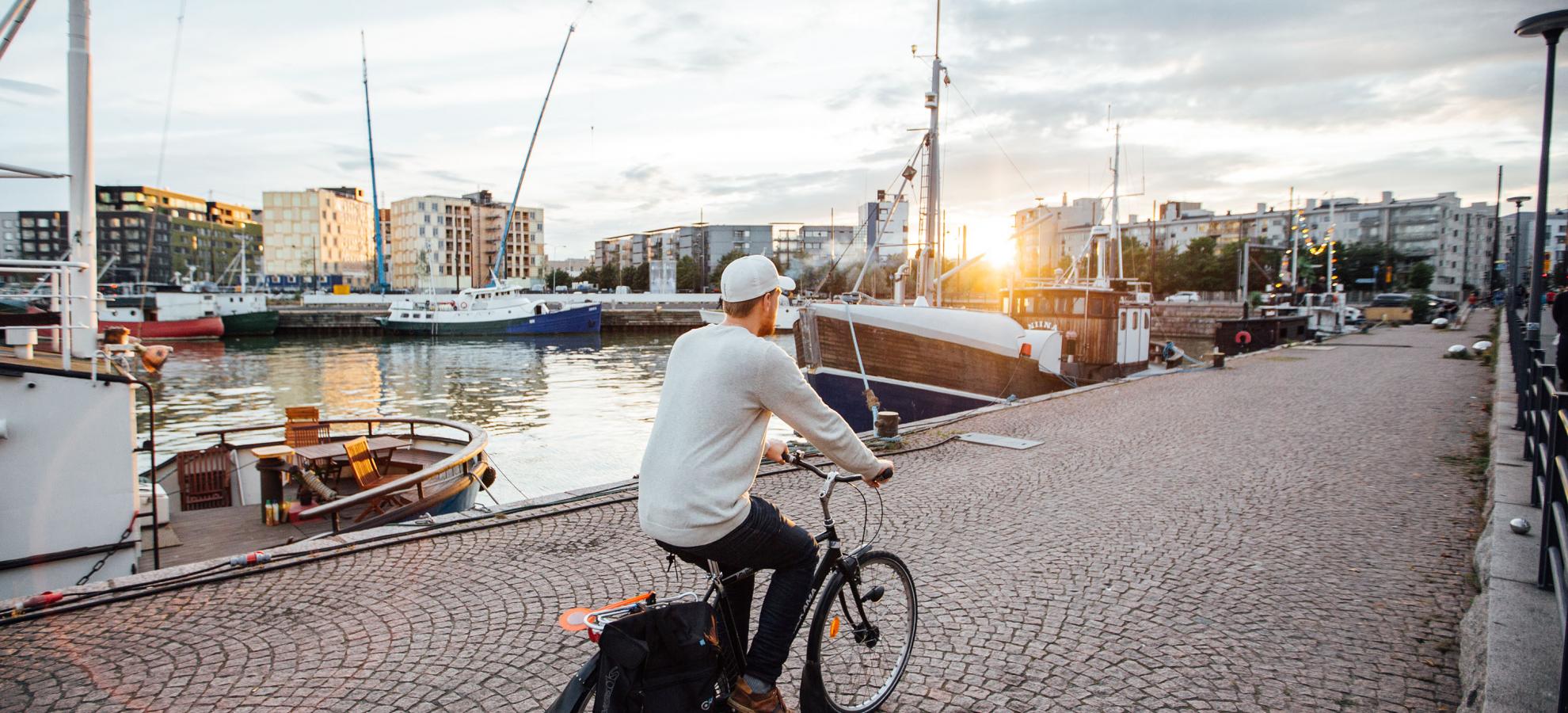 A man dressed in a white sweatshirt and cap, cycles away from the camera past docked boats, alongside west harbour as the sun sets. Apartment blocks are sat on the horizon on the edge of the harbour. 