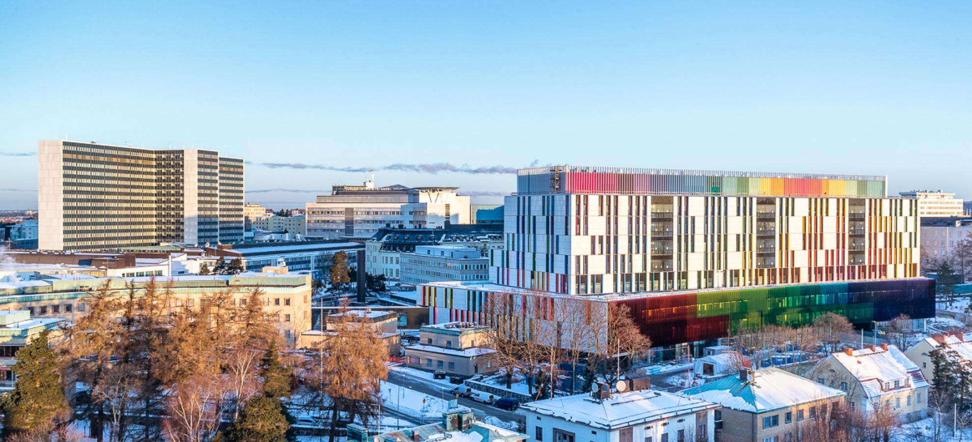 A view taken from a height of Helsinki Children's Hospital in Meilahti, a colourful building surrounded by other houses, offices and hospital buildings. There is snow on the ground. 