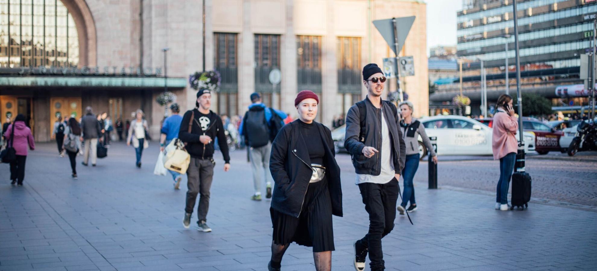 A man and woman, both dressed in black and grey and wearing beanies, are walking away from the Elielinaukio entrance of Helsinki Central railway station, which can be seen in the background. A small throng of people are walking on the pavement behind them in the distance.