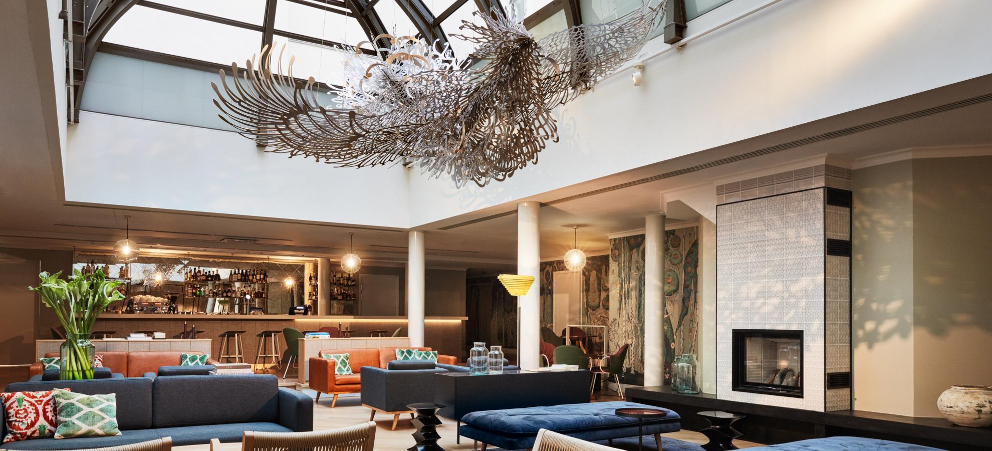 Hotel St.George Wintergarden, a large seated area filled with luxurious sofas and chairs underneath a skylight that stretches almost the whole length of the room. Pekka Jylhä's "Learning to Fly" sculpture hangs from the ceiling of the skylight..
